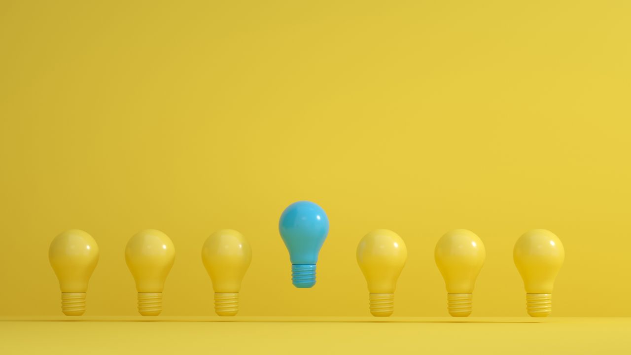 About A standout blue light bulb among several yellow bulbs on a yellow background, symbolizing innovation.